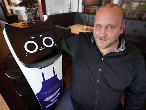 Ein Mitarbeiter serviert im Hafenrestaurant in Grömitz mit Servierroboter „Bella“ an einem Tisch Getränke. Foto: Marcus Brandt/dpa