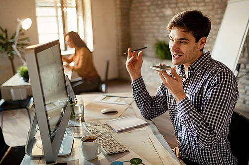 Lächelnder Freiberufler, der über den Lautsprecher des Mobiltelefons kommuniziert, während er Berichte auf einem Computer im Büro liest