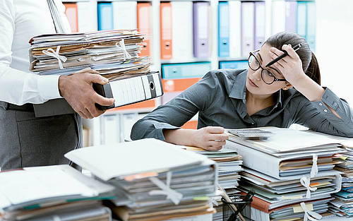 Gestresste Bürokraft vor einem Berg aus Dokumenten