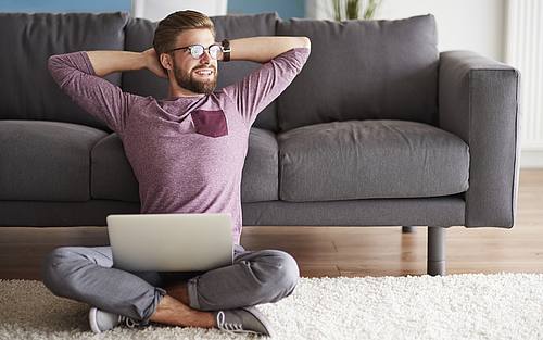 Ein Mann sitzt mit einem Laptop auf dem Boden vor einem Sofa.