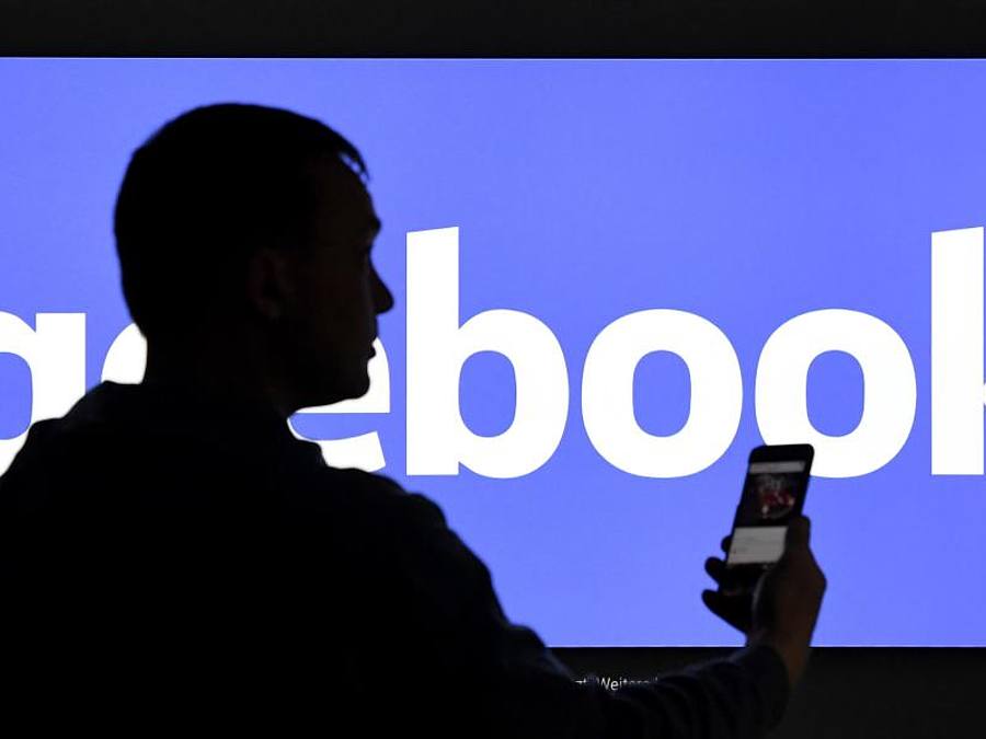 Ein Mann mit Smartphone steht vor einem Monitor mit dem Logo von Facebook. Foto: Carsten Rehder/dpa